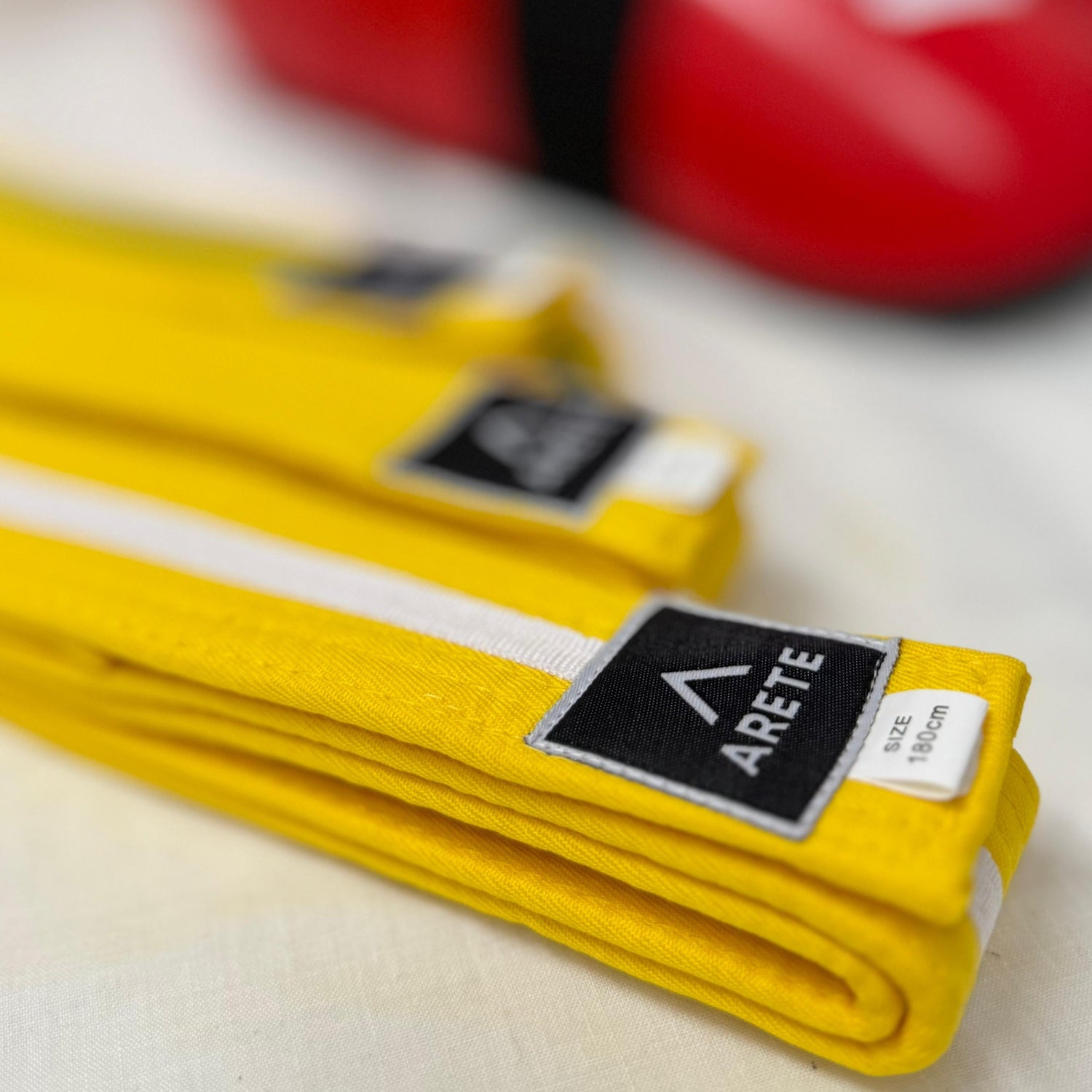 Yellow martial arts belt with Arete branding in the foreground, blurred red martial arts shoes in the background.