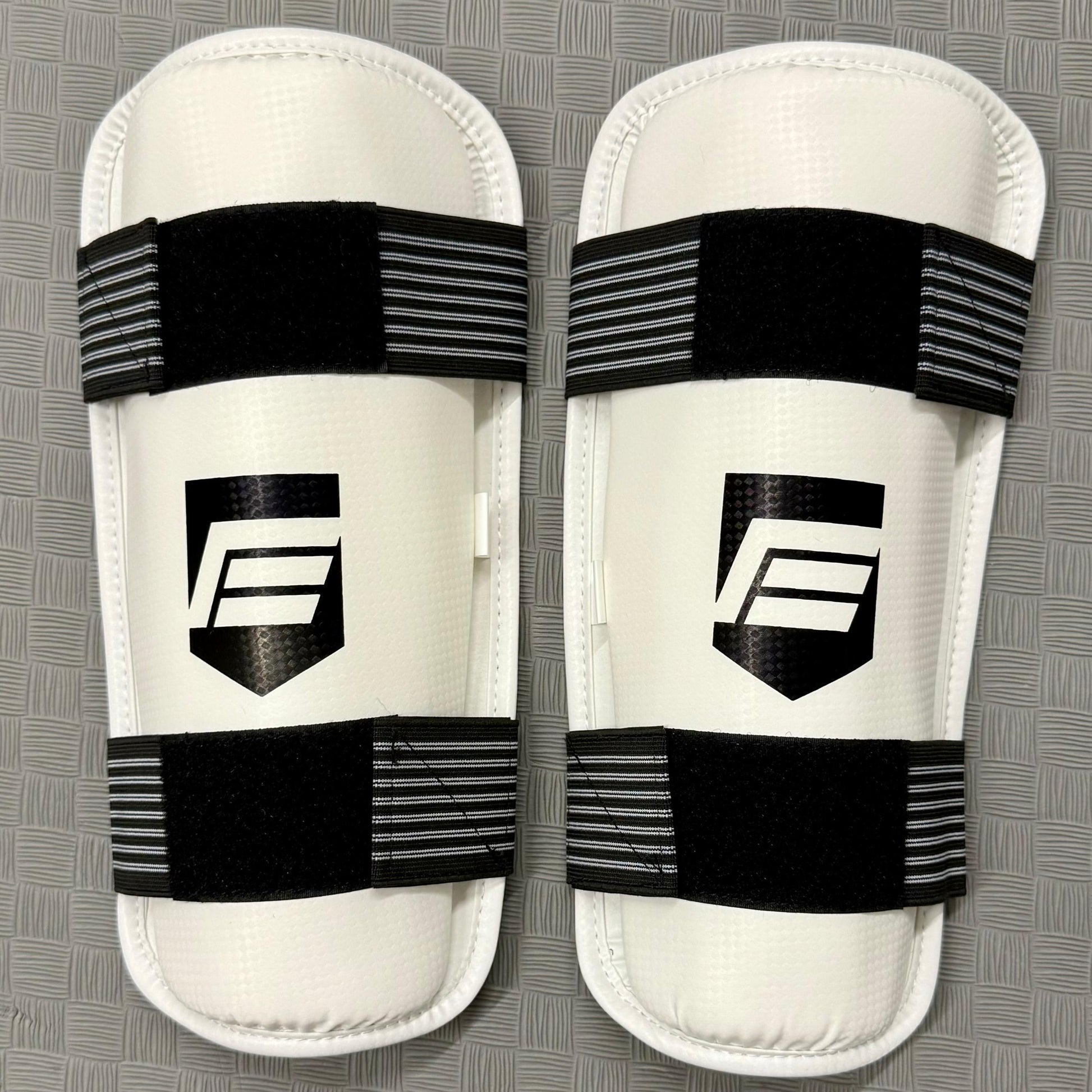 Pair of white shin guards with black straps on a grey background